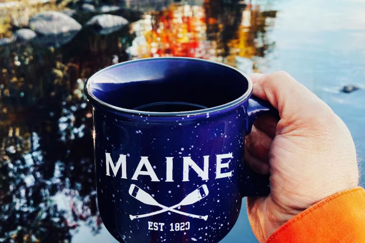 Hand holding a Maine mug over a still lake